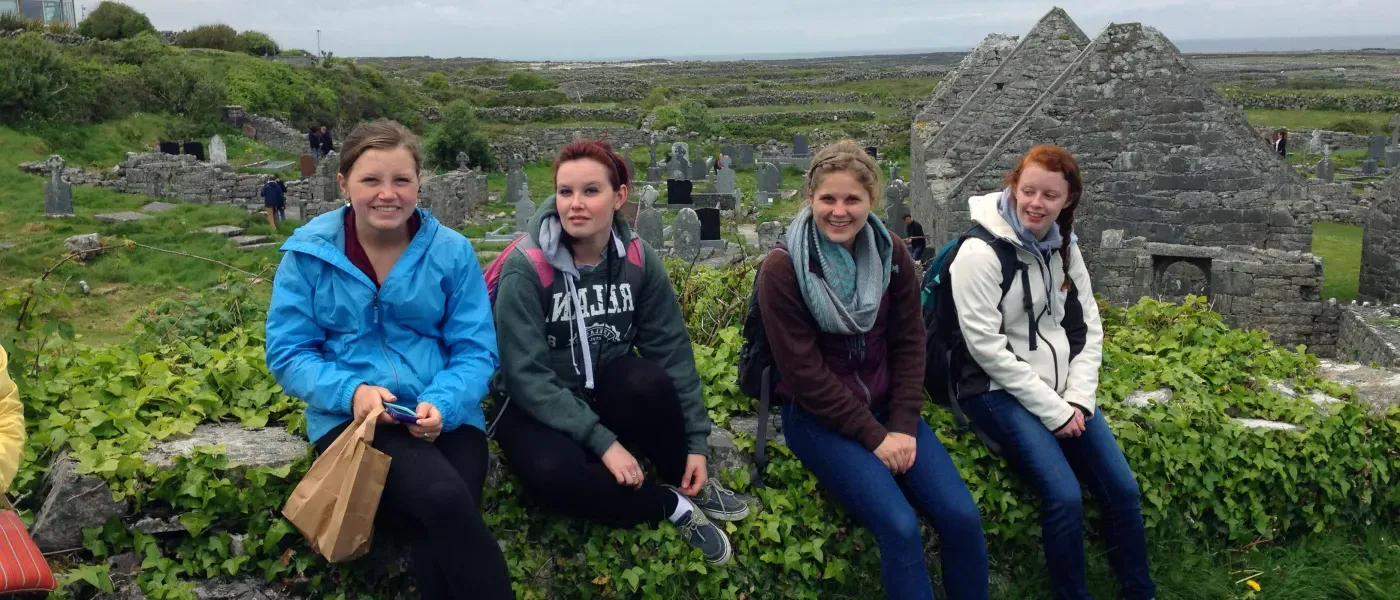 A group of U N E students sit on a stone wall at a rolling hillside cemetery in Ireland